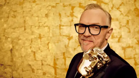 BBC/Bafta/Sane Seven Greg Davies posing with a gold Bafta mask trophy in front of a golden backdrop