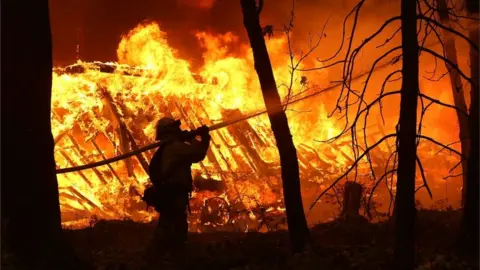 Getty Images A Cal Fire firefighter sprays water on a home next to a burning home as the Camp Fire moves through the area