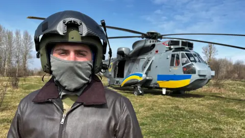 BBC Ukrainian pilot Vasil, pictured with his face covered in front of a Sea King helicopter donated by the UK and painted with the blue and yellow stripes of the Ukrainian flag.