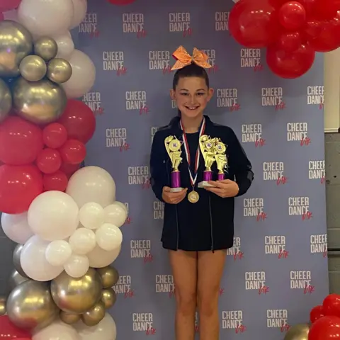 Amanda Axiak A young girl wearing an oversized black sweatshirt. She has her hair tied up with a large orange bow at the front. She has a medal around her neck and is holding three troophies. She is stood in front of a pink and blue background with the words "Cheer Dance" on it repeatedly. There ar epink, white and gold balloons saurrounding her and she is smiling and looking at the camera.