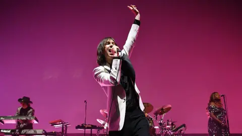 Getty Images Primal Scream performing on stage. Singer Bobby Giillespie has shoulder-length, dark hair and is wearing a light suit jacket over dark clothes. He is singing into a mic with his arm raised in the air. 