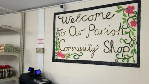 A 'Welcome to our parish community shop' sign which has a black border and green and pink flowers around it. There is a black till sitting on the bench and in the background you can see coffee pots and tinfoil sitting on shop shelves.