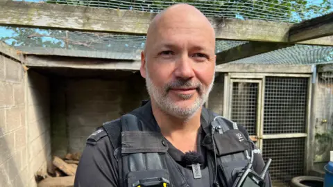 Detective Constable Aaron Flint stands inside a badger enclosure. He has a bald head and short grey beard. He is wearing a black police officer's uniform and is standing in front of an animal enclosure.