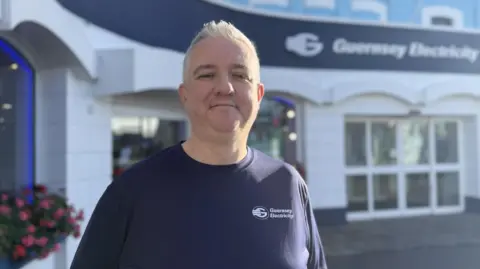 Gareth Wordingham facing the camera, standing outside a building which says "Guernsey Electricity" on it. He has short grey hair and is wearing a T-shirt with the Guernsey Electricity logo on it.