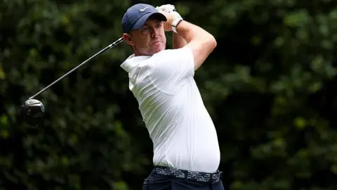 Rory McIlroy at the Truist Championship, wearing a white golf shirt and blue cap, swinging a metal wood