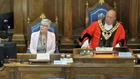 BBC/Lucy Ashton A man with a bald head is wearing a bright red and black robe, with a white frill and large gold chain. He is sitting next to a woman with short grey hair and glasses wearing a pale pink blazer and grey striped shirt. They are sitting at a large wooden desk in a council chamber