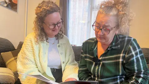 Suzanna and Sarah looking at a Christians Against Poverty debt pamphlet. Suzanna is wearing a textured light yellow cardigan and Sarah is wearing a green, black and white checker shirt sitting on a ribbed grey sofa.