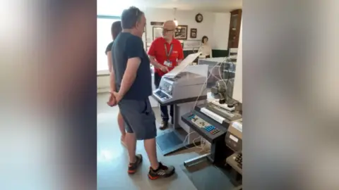 Amberley Museum A man in a red polo shirt demonstrating to two people how a large grey Telex machine works
