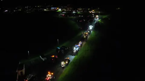 Lit up tractors drive down a lane of a village in the dark, seen from above in a drone shot.