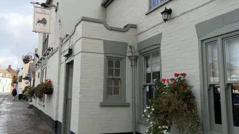 The exterior of the Black Bull pub. The walls are white and the window frames are painted a sage green. Flowers cascade from baskets, which are mounted to the walls.