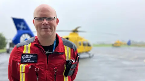 Dr Tim Smith wearing a red and yellow uniform in front of the air ambulance