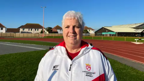 Jean Cross, Vice-President of the Commonwealth Games Association of Jersey, at FB Fields Athletics Track, wearing a Team Jersey branded sports jacket 
