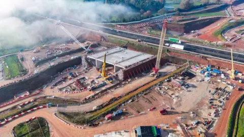 A large construction site, with a tunnel being built next to a duel carriageway, with a number of crane and construction vehicles near by.