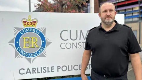 Insp Andy Leather outside Carlisle Police Station. He is a large man with short bangs, graying hair and a full beard. He is wearing a black t-shirt and pants. Behind him is a large sign for Carlisle Police Station, featuring the Cumbria Constabulary logo. A small part of the building is out of focus behind him.