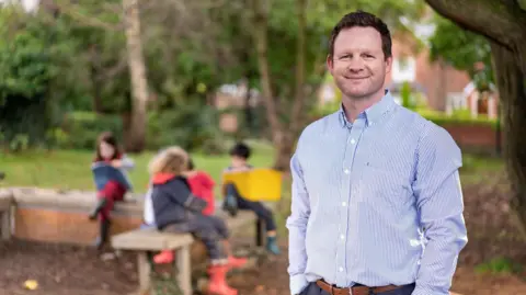 Handout a man in a shirt smiling with his hands in his pockets. Behind him, out of focus, a few children in waterproof clothing and boots sit on a bench and write or draw in large notebooks. The photo was taken outside and some trees are visible in the background