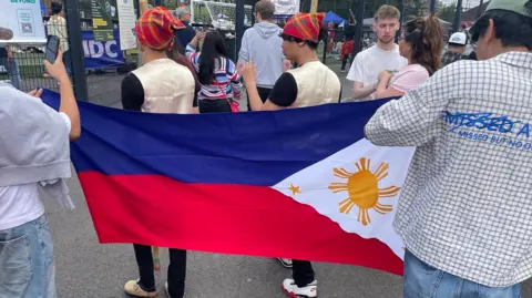 Two men hold a flag of the Philippines. It has a blue bar and a red bar underneath, a white triangle pierces the colours from the right with a yellow sun in the white part. Two men dressed in gold waistcoats and black t-shirts walk on the other side of the flag. The man holding the flag on the right wears a white and brown chequered shirt and green hat with blue jeans. The other man has on blue jeans and a white t-shirt, with a grey hoodie round his neck.