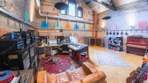 Steve Macarthur The inside of the recording studio in a brick-lined spacious room with various instruments, including a piano, keyboard and guitars and and sizable recording equipment