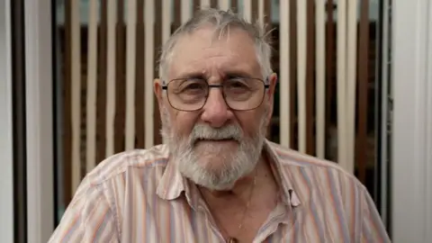 Close-up of Alan, 83, who has closely cropped grey hair and beard and wears large black-rimmed glasses.