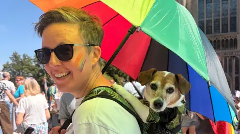 Harriet Morter/BBC A woman with short cropped hair holding a rainbow umbrella with a dog inside a backpack on her back. She has a pair of sunglasses on her eyes and both her and the dog are looking at the camera.