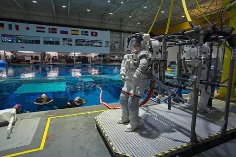 Kevin Church/BBC Image of Rosemary Coogan taken from her left side as she waits by the side of the training pool at Johnson Space Centre. There is a large red "umbilical hose" coming out of the large white pack on her back and going into the water in the deep blue swimming pool to her right. Mock ups of the space station can be seen below the surface. There are two divers in the water. On the other side of the pool is a white building containing the control centre with windows overlooking the pool. Above the windows are some the flags of the countries involved in the space station. 