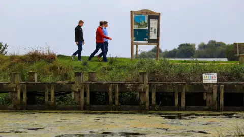 PA Media Three people walking along the bank of a waterway. The water has green algae on the surface