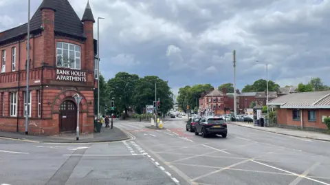 Two cars waiting at a set of traffic lights. The road continues ahead and has a filter to a road to the left. To the left of the cars is a red-brick building with the words 'Bank House Apartments' written on front.