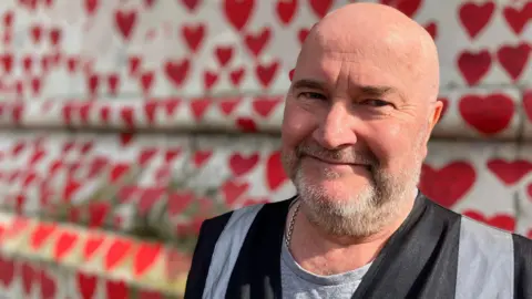 A bald-headed man with silver beard smiles kindly towards the camera against the backdrop of a wall painted with red hearts