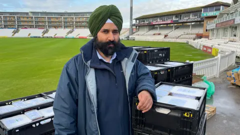 Raj Singh wearing a blue coat and green turban is stood in the middle of the picture, with his arm resting on a pile of crates full of food to be packed into vans. In the background is the pitch and stands of Somerset County Cricket Club.