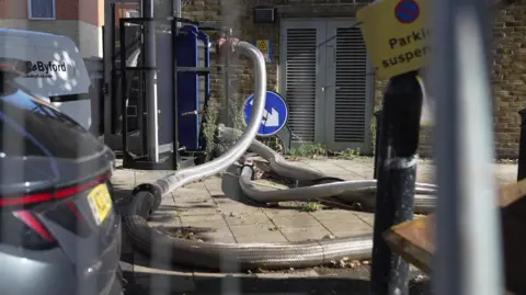 Pipes running along a pavement with cars parked near-by