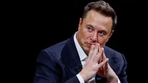 Elon Musk on a stage with his hands tented in front of his mouth as he listens to someone next to him.