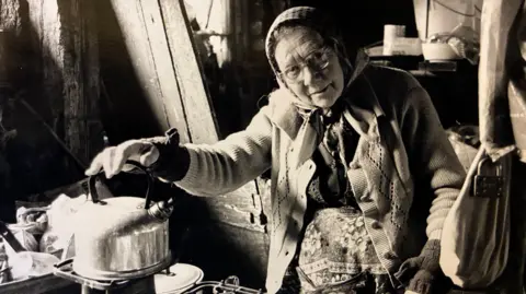 Submitted An elderly woman wearing a cardigan, headscarf, gloves and glasses looks directly at the camera. She is next to timbers from an unfinished house and has her right hand on a kettle which appears to be on some kind of stove. 