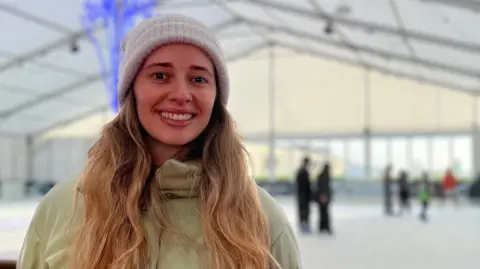 Anabela is wearing a light green winter coat and a knitted hat is standing in front of an indoor ice-skating rink. The rink is covered by a large white tent structure with beams and lights overhead. In the background, several people are skating, and there are blue decorative lights shaped like trees.