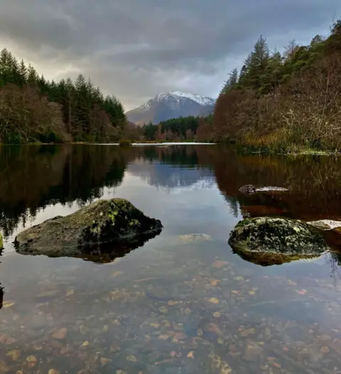 Morag MacNeil A mountain is framed by forest on both sides of the photo and a loch in the foreground