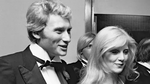 Getty Images A black and white photo taken in Paris, 1975 of Johnny Hallyday smiling while wearing a black suit with bowtie and flower on his lapel accompanied his wife Sylvie Tartan who has blonde hair and is also smiling.