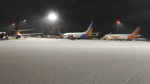 Charlie Haskayne Three planes on snow-covered tarmac at Bristol Airport. A set of vehicle tracks can be seen in the foreground, but otherwise the snow is completely undisturbed.