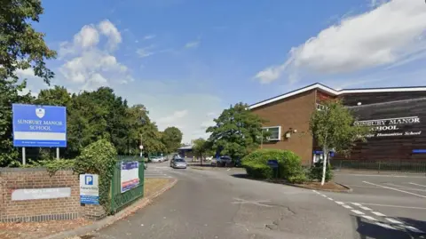 Google The outside of Sunbury Manor School seen in a Google Street View image. There is a blue sign on the left with the school name on and the buildings can be seen on the right on the other side of a road running in to the school.