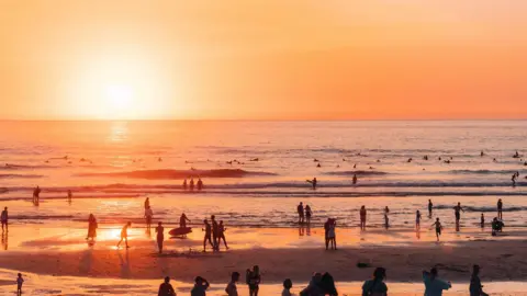Darina Fistral Crowds are walking in the sea holding surfboards as waves crash around them. The sun is golden and setting in the distance. 
