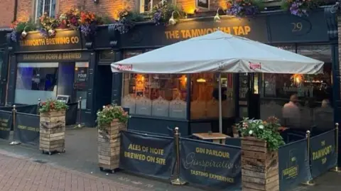 Camra There is a big beer garden umbrella outside and the words The Tamworth Tap are in yellow near the entrance. Plants are also near the entrance.
