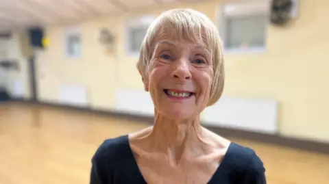 Aimee Dexter/BBC Barbara Wraith is standing in a hall. She has short blonde hair and is wearing pink lipstick and a black V-neck T-shirt.