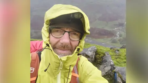 A man taking a selfie up a large hill, with a view looking down it in the background. He has a black cap on with black glasses and is wearing a neon yellow waterproof jacket with the hood up, and an orange rucksack