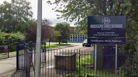 Google The entrance to a school, with a sign saying Biddulph High School. There are railings, and a blue coloured building can be seen behind
