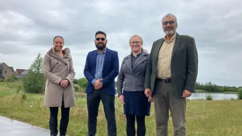 JOANNA TAYLOR/BBC Four people standing in a line on a path. They are smiling at the camera. From left to right, they are Kate Nation from the Turtle Dove social enterprise, Atteq Nasir from Northstowe Muslims, the Reverend Beth Cope from the Northstowe Church Network and Jawad Nawash from Northstowe Muslims. Behind them is grass and a lake, Bug Hunter Waters. 