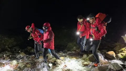 Keswick Mountain Rescue Four people in red coats walk through a stream in the dark