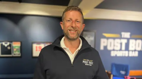 Max Rauber is wearing a dark blue jacket and is behind a backdrop of football t-shirts. 