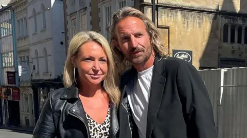 Nigel Adams wearing a white T-shirt and black blazer. He has long blonde hair and a dark beard. He is standing beside his fiancee Louise Payne, who has blonde hair and is wearing large hoop earrings, a cheetah print blouse and a black leather jacket.