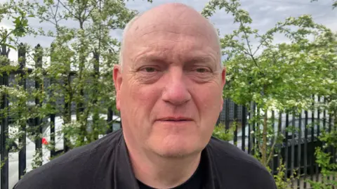 A man with short grey hair at the backs and sides of his head stares at the camera. He wears a black jacket and black T-shirt as he stands in front of a black metal fence with green trees in front of it. 