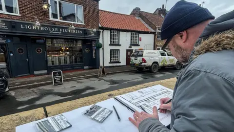 Jordan Lea A man in a grey thick coat and navy beanie is drawing a building in a large sketchbook with pencils next to him. A small road is in front of him with a building opposite which has a black exterior and reads "Lighthouse Fisheries" in white lettering across its front.