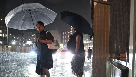 Two women with umbrellas waiting in the rain at night