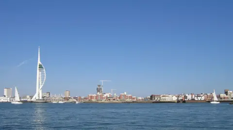 Getty Images The Portsmouth skyline, taken from out to sea. There's the tall white Spinnaker Tower on the left, some shorter brick buildings in the middle with cranes behind them, and some white buildings on the right.