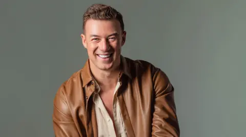 A light brown haired man scratched, wearing a brown leather jacket and a light brown shirt with the top buttons cancelled. He smiled on the camera.